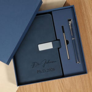 May include: A navy blue gift set containing a notebook and pen. The notebook has a silver clasp and the name "Dr. Johnson Ph.D.2026" engraved on the cover. The pen is silver and in clear packaging. The set is presented in a navy blue box.