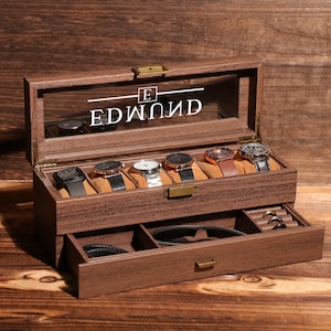 May include: A wooden watch display case with a glass top, featuring the name "EDMUND" in white lettering. The case holds six watches on tan cushions and has a lower drawer for accessories. The case is brown.