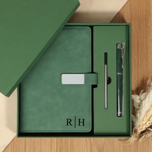 Puede incluir: Un juego de diario y bolígrafo verde en una caja de regalo. El diario tiene un cierre plateado y las iniciales "R | H" impresas en la portada. El bolígrafo y una recarga están incluidos. La caja es verde.