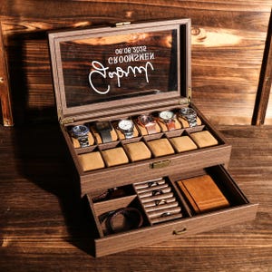 May include: A wooden watch box with a glass top, displaying several watches on brown cushions. The box has a drawer with compartments for jewelry and small accessories. The text "groommen" is visible on the glass.