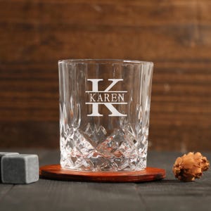 Puede incluir: Un vaso de whisky transparente grabado con un patrón de corte de diamante. El vaso presenta el nombre "KAREN" con la letra "K" arriba y abajo. El vaso se encuentra sobre un pequeño posavasos redondo de madera. Junto al vaso hay tres cubitos de hielo cuadrados grises y un objeto decorativo.