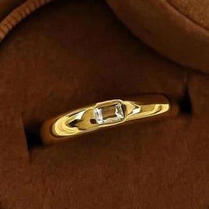 Puede incluir: Un anillo de oro con una gema rectangular y transparente. El anillo se muestra dentro de un joyero marrón forrado de terciopelo. El anillo tiene un acabado liso y pulido y un diseño clásico.