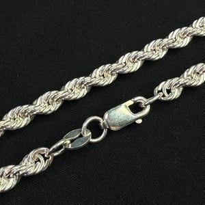 May include: Silver-colored rope chain necklace with a lobster clasp. The chain is made of twisted metal links, creating a rope-like texture. The clasp is attached to the chain with a small ring. The background is black.