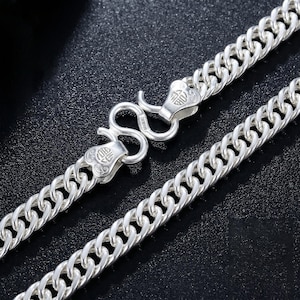 May include: A silver chain necklace featuring interlocking curb links and an S-hook clasp. The clasp ends are shaped like stylized hearts and are engraved with a small circular symbol containing Chinese characters, providing a secure closure.