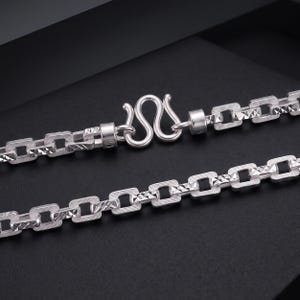 May include: A silver-toned chain necklace featuring rectangular links with a textured, diamond-cut finish. The chain is secured with an S-shaped hook clasp, and each link displays a repeating pattern of small, reflective geometric facets.