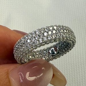 May include: A silver ring with multiple rows of small, clear gemstones. The ring is held by a person, and the gemstones sparkle in the light. The ring appears to be a band style, with the gemstones covering the entire surface.