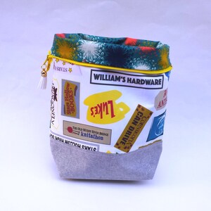 May include: A white fabric drawstring bag with a yellow top and a gray bottom. The bag is decorated with colorful patches that say "Stars Hollow", "Luke's", "William's Hardware", "The Old Muddy River Bridge Knitathon", "Cornucopia Can Drive", and "Ante".
