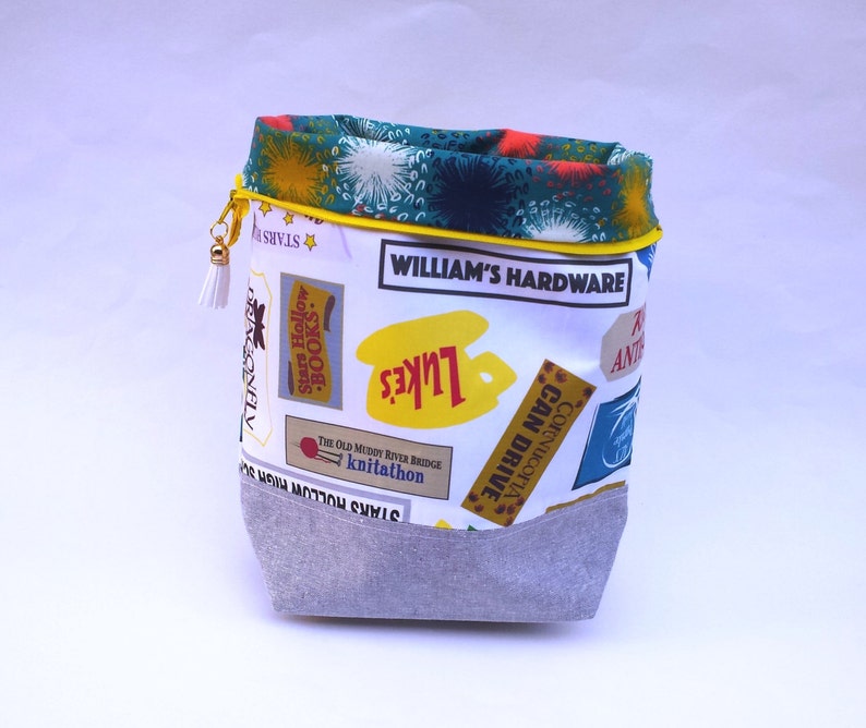 May include: A white fabric drawstring bag with a yellow top and a gray bottom. The bag is decorated with colorful patches that say "Stars Hollow", "Luke's", "William's Hardware", "The Old Muddy River Bridge Knitathon", "Cornucopia Can Drive", and "Ante".