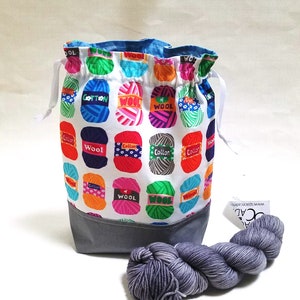 May include: A white drawstring bag with a colorful print of yarn balls. The print includes the words "Wool" and "Cotton". The bag has a gray bottom and a white drawstring. A skein of gray yarn is in the foreground.