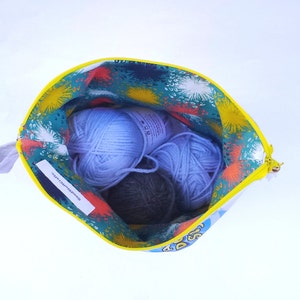 May include: A yellow zippered pouch with a colorful floral print and the text "Good things take time" inside. The pouch is open and contains three balls of yarn: two light blue and one dark gray.