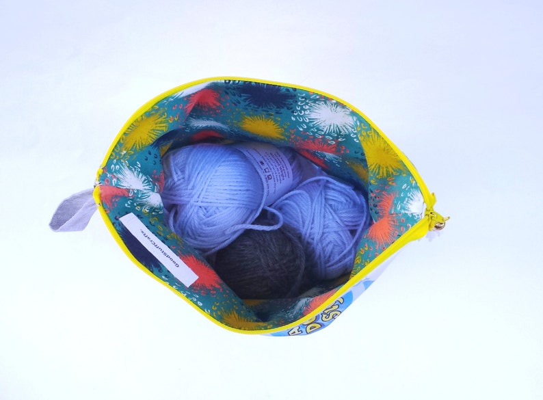 May include: A yellow zippered pouch with a colorful floral print and the text "Good things take time" inside. The pouch is open and contains three balls of yarn: two light blue and one dark gray.