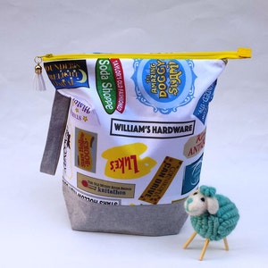 May include: A white zippered pouch with a gray bottom. The pouch is decorated with a variety of colorful logos and text, including "Soda Shoppe", "Taylor's Old Fashioned", "The Amazing Doggy Sam", and "William's Hardware". A small blue and white yarn sheep is standing in front of the pouch.