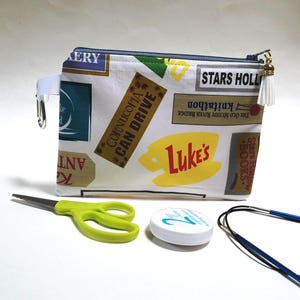 May include: A white zippered pouch with a colorful pattern of vintage-style signs and text, including "Cornucopia Can Drive", "Luke's", and "Stars Hollow". The pouch has a gold zipper pull with a white tassel.