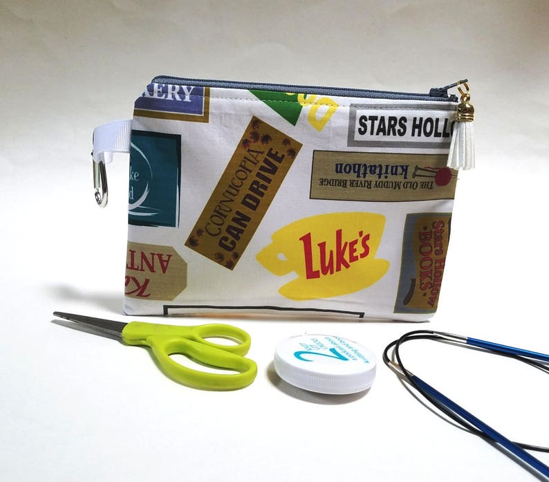 May include: A white zippered pouch with a colorful pattern of vintage-style signs and text, including "Cornucopia Can Drive", "Luke's", and "Stars Hollow". The pouch has a gold zipper pull with a white tassel.
