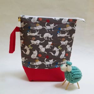 May include: A gray zippered pouch with a red bottom. The pouch is decorated with a pattern of white cats playing with balls of yarn. A small teal blue sheep figurine is in the foreground.