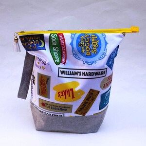 May include: A white zippered pouch with a gray bottom. The pouch is decorated with colorful patches featuring various text, including "Soda Shoppe", "William's Hardware", "The Amazing Doggy Swim", and "Stars Hollow".