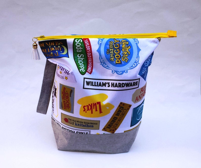 May include: A white zippered pouch with a gray bottom. The pouch is decorated with colorful patches featuring various text, including "Soda Shoppe", "William's Hardware", "The Amazing Doggy Swim", and "Stars Hollow".