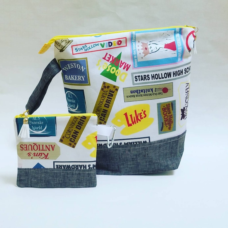 May include: Two fabric bags with a Stars Hollow theme. The larger bag is white with a blue denim bottom and features various Stars Hollow businesses and locations printed on it. The smaller bag is white with a blue denim bottom and features a Stars Hollow business and location printed on it.