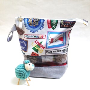 May include: A gray zippered pouch with a Stars Hollow theme. The pouch is filled with yarn balls and has a clear plastic window. The pouch is sitting on a white surface.