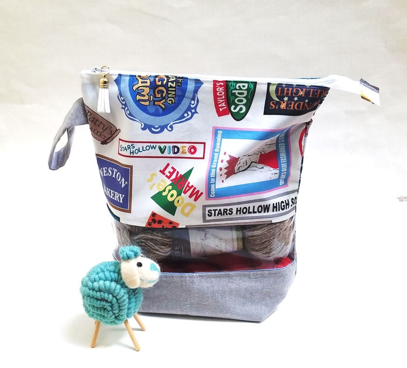 May include: A gray zippered pouch with a Stars Hollow theme. The pouch is filled with yarn balls and has a clear plastic window. The pouch is sitting on a white surface.