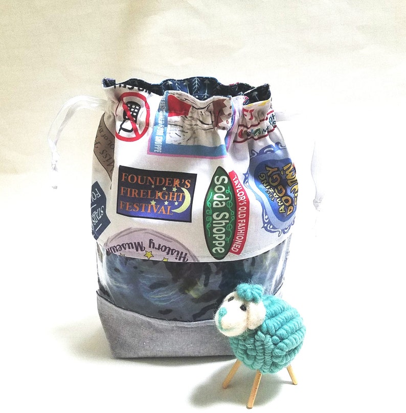 May include: A white drawstring bag with a blue and white fabric lining. The bag is decorated with colorful patches featuring text like "Soda Shoppe", "Founder's Firelight Festival", and "Taylor's Old Fashioned". A small blue crocheted sheep with wooden legs sits in front of the bag.