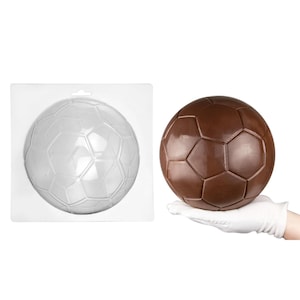 May include: A brown chocolate soccer ball held in a gloved hand, next to a clear plastic mold in the shape of a soccer ball. The chocolate ball has a glossy finish and is made of many pentagonal and hexagonal shapes.