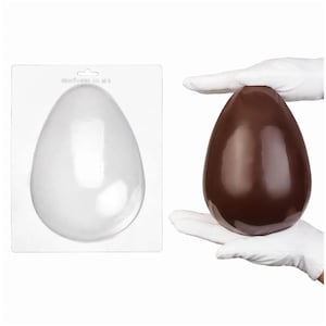 Könnte beinhalten: Eine transparente Eierform aus Kunststoff neben einem großen, massiven Schokoladenei. Das Schokoladenei wird in behandschuhten Händen gehalten. Die Form ist eine klare, eiförmige Form, die an einer weißen Rückseite befestigt ist.