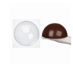 Puede incluir: Un molde de plástico transparente y redondo junto a un chocolate marrón oscuro y semiesférico. El chocolate está sostenido por una mano enguantada. El molde es cuadrado con una hendidura circular.