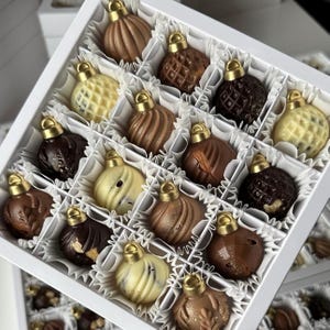 Puede incluir: Una caja blanca llena de adornos de chocolate surtidos. Cada adorno tiene una tapa dorada y está elaborado en varios tonos de chocolate marrón y blanco. Los chocolates están dispuestos en compartimentos individuales.