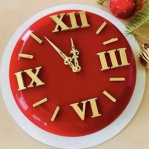 Può includere: Un orologio rosso con numeri romani dorati e lancette dorate. Il quadrante è lucido e posto su uno sfondo bianco. L'orologio indica che sono quasi le 12.