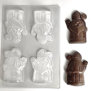May include: A clear plastic rectangular mold featuring four cavities shaped like Saint Nicholas, accompanied by two finished brown chocolate figures of Saint Nicholas, each depicting a bearded figure wearing a tall miter hat and holding a staff.