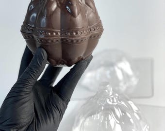 May include: A brown, molded chocolate ornament with a detailed, embossed floral and beaded pattern. The chocolate is shaped like a rounded, segmented sphere with a small top protrusion. Beside it sits a clear, empty plastic mold of the same shape.