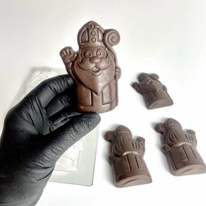 May include: A molded milk chocolate figure of Saint Nicholas wearing a mitre and holding a staff, shown in a brown finish. The figure features a smiling face with a beard and is presented alongside three identical chocolate pieces and a plastic mold.