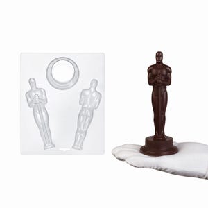 May include: A chocolate-colored Oscar statuette held in a hand, next to a clear plastic mold with two statuette impressions. The statuette is standing on a round base and holding a sword. The mold has a circular opening at the top.