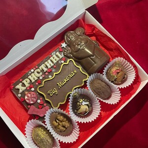 May include: A white gift box containing a large rectangular milk chocolate bar with the text "ХРИСТИНІ" and "Від Миколая" in gold, a molded chocolate figure of Saint Nicholas, and five assorted chocolate truffles with nuts and gold accents.