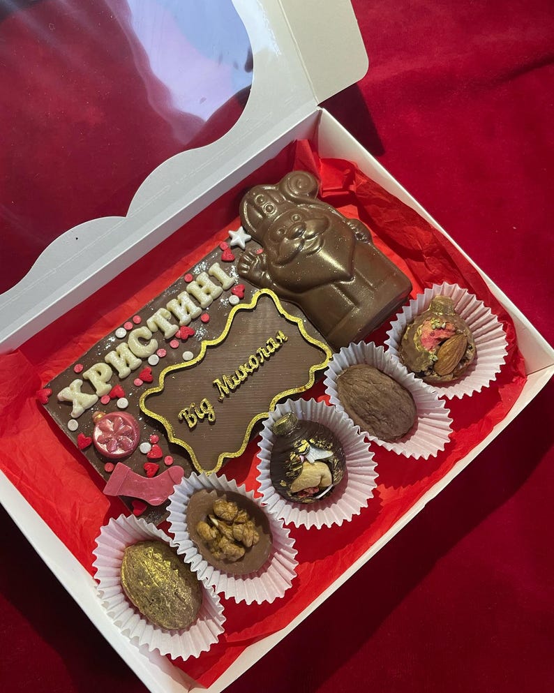 May include: A white gift box containing a large rectangular milk chocolate bar with the text "ХРИСТИНІ" and "Від Миколая" in gold, a molded chocolate figure of Saint Nicholas, and five assorted chocolate truffles with nuts and gold accents.