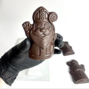 May include: A molded dark chocolate figure of Saint Nicholas wearing a mitre and holding a crosier. The figure features a smiling face with a mustache and a raised hand. Two additional identical chocolate figures are visible in the background.