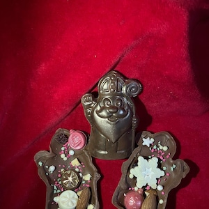 May include: A dark chocolate figure of Saint Nicholas positioned between two dark chocolate mittens. The mittens are decorated with white chocolate snowflakes, pink and white sugar pearls, dried fruit pieces, and whole almonds.