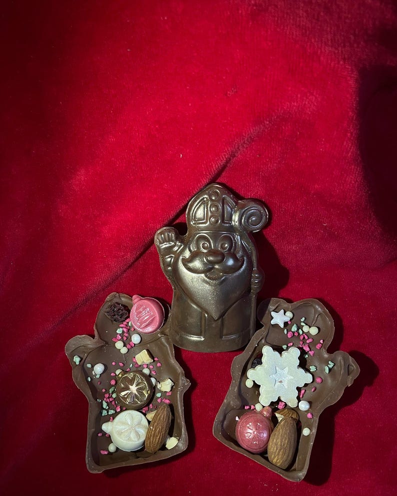 May include: A dark chocolate figure of Saint Nicholas positioned between two dark chocolate mittens. The mittens are decorated with white chocolate snowflakes, pink and white sugar pearls, dried fruit pieces, and whole almonds.