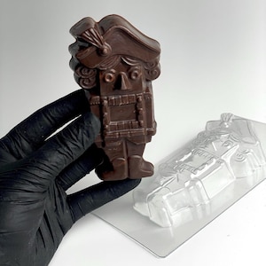 Könnte beinhalten: Eine dunkelbraune Schokoladen-Nussknacker-Figur, die von einer behandschuhten Hand gehalten wird, neben einer transparenten Kunststoffform. Der Nussknacker hat ein detailliertes Design, einschließlich Hut, lockigem Haar und Uniform. Die Form hat die Form des Nussknackers.