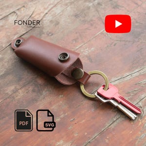 Puede incluir: Un llavero de cuero marrón con un llavero de latón y dos llaves. Las llaves son rojas y plateadas. El llavero tiene un cierre a presión. La palabra "FONDER" está impresa en la parte superior izquierda.