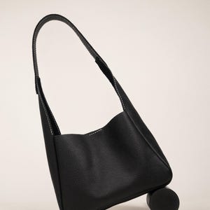 May include: A black leather shoulder bag with a curved handle and white stitching. The bag is resting on a small, black cylindrical object. The bag's design is minimalist and modern, suitable for everyday use.