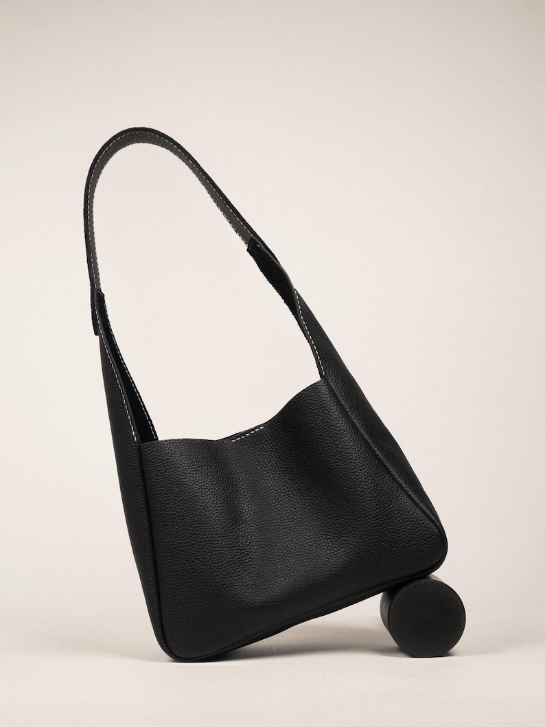 May include: A black leather shoulder bag with a curved handle and white stitching. The bag is resting on a small, black cylindrical object. The bag's design is minimalist and modern, suitable for everyday use.