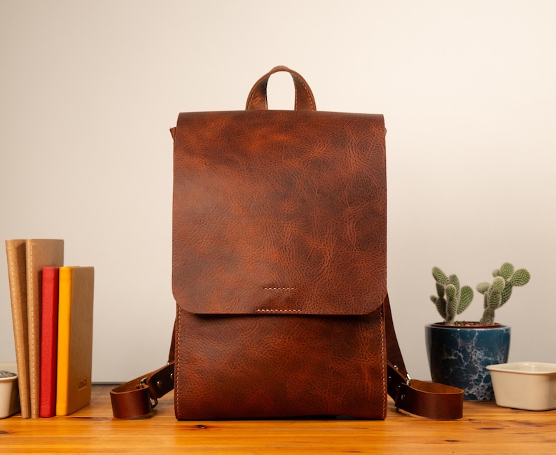 May include: A brown leather backpack with a flap closure and adjustable straps. The bag has a top handle and is sitting on a wooden surface. A stack of books and a potted cactus are in the background.