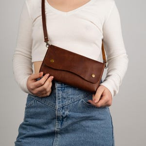 Peut inclure: Un sac bandouli&egrave;re en cuir marron avec une fermeture &agrave; rabat et deux rivets couleur laiton. Le sac est dot&eacute; d'une longue bandouli&egrave;re r&eacute;glable. Le sac est tenu par une personne portant un t-shirt blanc &agrave; manches longues et une jupe en jean.