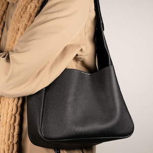May include: A black leather shoulder bag with a textured surface. The bag has a rectangular shape with a rounded bottom and a long, black strap. White stitching accents the edges of the bag.
