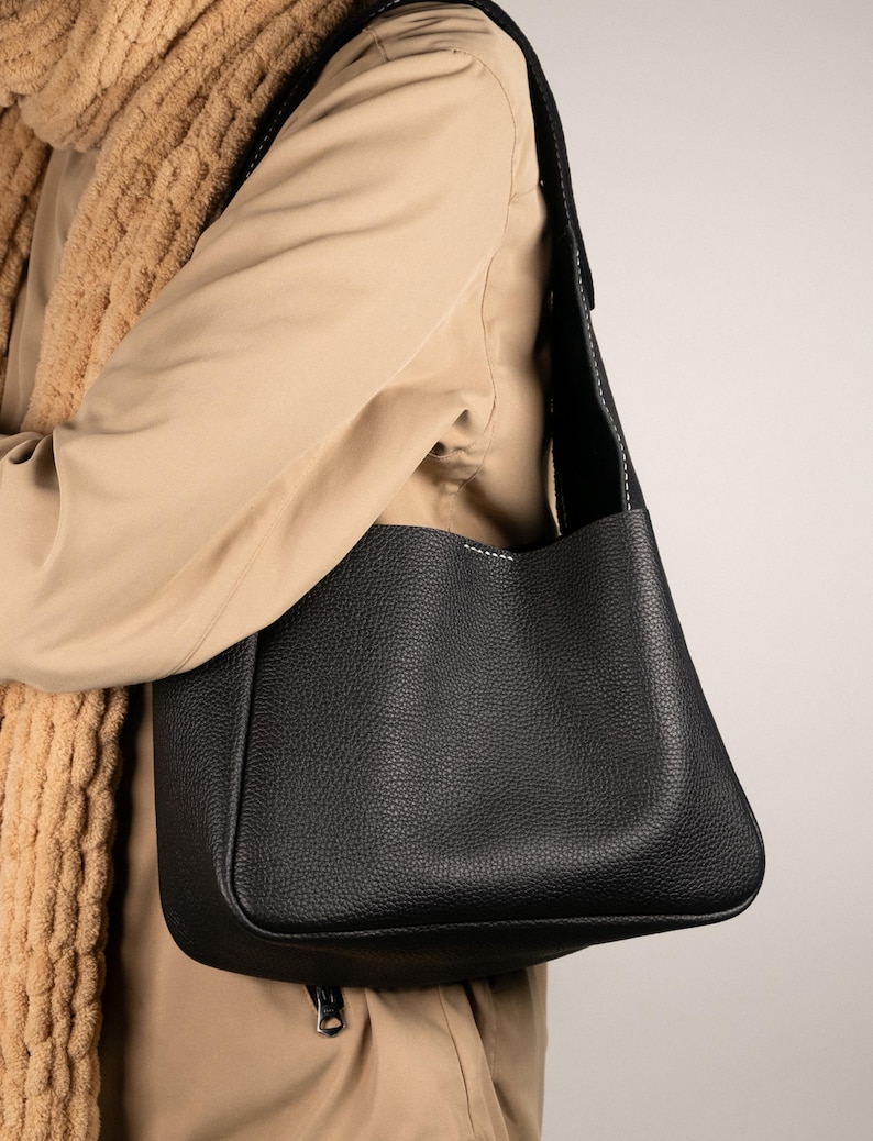 May include: A black leather shoulder bag with a textured surface. The bag has a rectangular shape with a rounded bottom and a long, black strap. White stitching accents the edges of the bag.