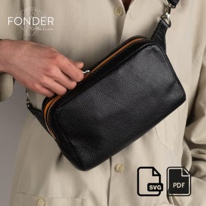 Op de afbeelding: Zwarte leren crossbodytas met een getextureerde afwerking en oranje ritssluiting. De tas heeft een rechthoekige vorm en een zilverkleurige ritssluiting. De tas is voorzien van de tekst "FONDER Atelier".