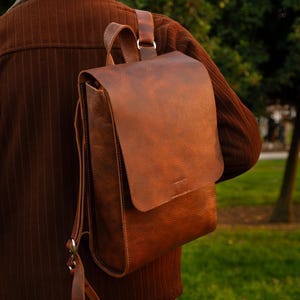 May include: A brown leather backpack with a flap closure and adjustable straps. The bag is worn over the shoulder, showcasing its textured surface and classic design. The backpack is a rich, warm brown color.