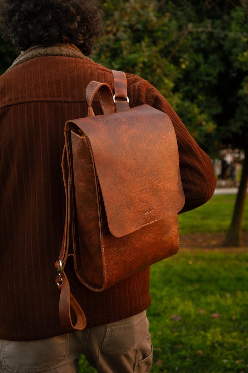 May include: A brown leather backpack with a flap closure and adjustable straps. The bag is worn over the shoulder, showcasing its textured surface and classic design. The backpack is a rich, warm brown color.